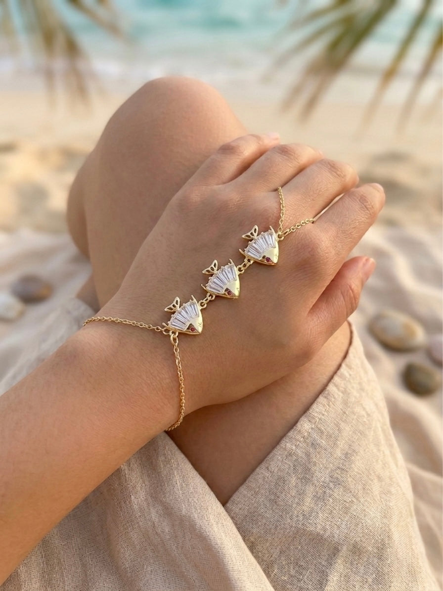 Hand wearing a gold bracelet with fish-shaped charms on a beach.