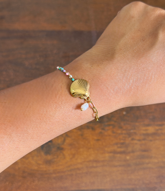 Gold bracelet with a shell charm on a wrist against a wooden background