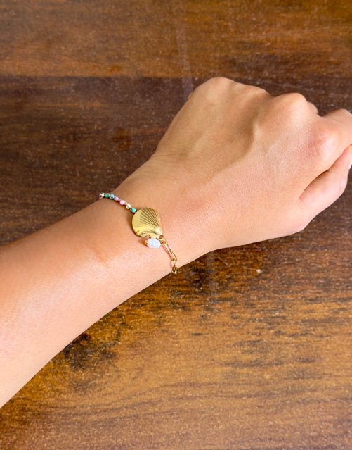 Hand wearing a gold bracelet with a shell charm on a wooden surface